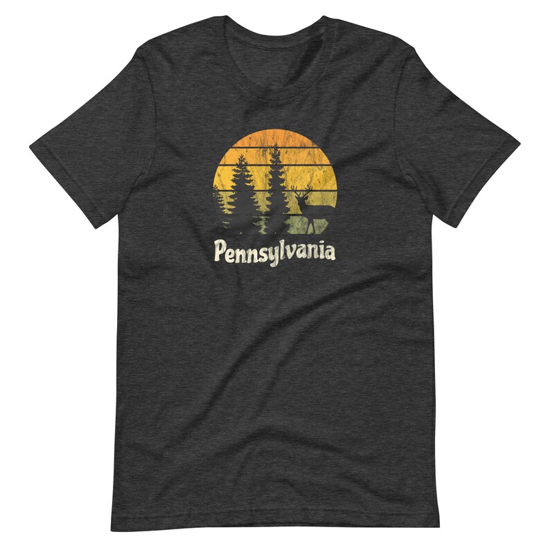 Op de afbeelding: Donkergrijs t-shirt met een vintage-stijl afbeelding van een zonsondergang, bomen en een hert. De tekst "Pennsylvania" is onder de afbeelding gedrukt.
