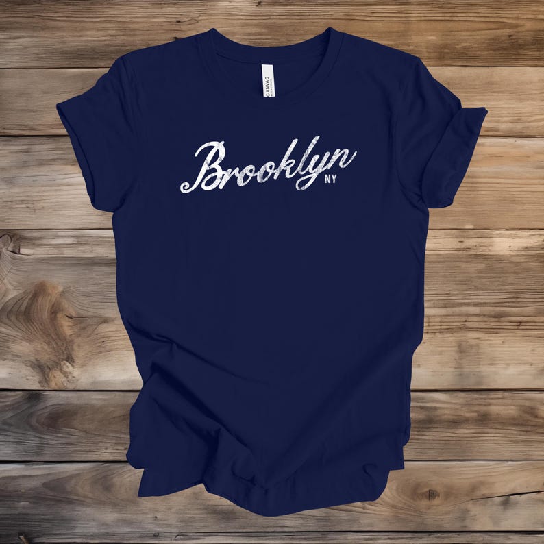 Op de afbeelding: Marineblauw t-shirt met het woord "Brooklyn" in witte scriptletter. De letters "NY" staan eronder. Het t-shirt wordt gepresenteerd op een houten ondergrond.
