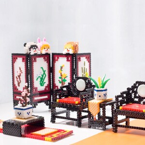 May include: A miniature living room set made of colourful beads, featuring a black beaded screen with floral designs, two black beaded chairs with red cushions, a small table with a yellow tablecloth, and a potted plant.