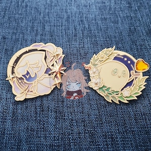 May include: Two gold-colored enamel pins with a whimsical design. The first pin features a crescent moon with stars and a white and blue floral design. The second pin features a crescent moon with a yellow star and a green and white floral design.