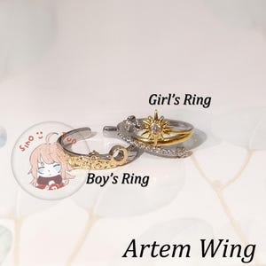 Puede incluir: Se muestran dos anillos y un botón. Un anillo es dorado y plateado con un diseño de sol. El otro anillo es dorado y plateado con un diseño decorativo. El botón tiene un personaje de dibujos animados. También se ve el texto "Girl's Ring", "Boy's Ring" y "Artem Wing".
