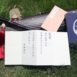 May include: A traditional Chinese instrument, a guqin, with a red tassel and a white card with Chinese characters. A pink book and a blue book are also visible. The text on the card reads "Send the volume, Lan's school is about to begin."