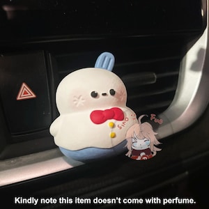 May include: A white, round, plush toy with a red bow tie and blue pants. The toy has a small, blue, pointed hat on its head. The toy is sitting in a car's air vent. The text "Sino" is visible on the toy's chest. The text "Fun" is visible in the background.