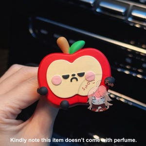 May include: A red apple-shaped car air freshener with a cute cartoon face. The apple has a frown and black eyes. The air freshener is made of soft, squishy material.