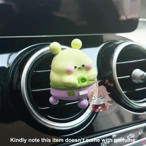 May include: A green and purple cartoon character air freshener hanging from a car's air vent. The character has a round body, big eyes, and a small mouth. The air freshener is labeled "Fun".