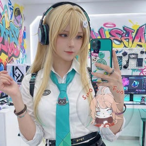 May include: A person wearing a white collared shirt, teal tie with a black cat pin, and black headphones. The person is holding a teal phone with stickers and a cartoon image. The background features graffiti art.