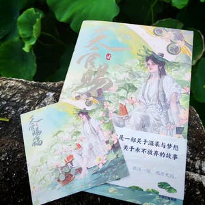 以下が含まれることがあります： 蓮の花が咲く池の近くに白いローブを着た人物のイラストが描かれた2冊の本。表紙には漢字とテキストが使用されています。背景は緑と青の柔らかいブレンドです。