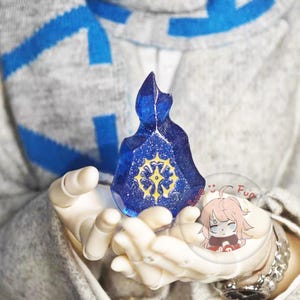 以下が含まれることがあります： 黄色いエンブレムとキラキラしたディテールが施された、青い宝石型のペンダント。ペンダントは両手に持たれています。背景は青いアクセントのグレーのセーターです。