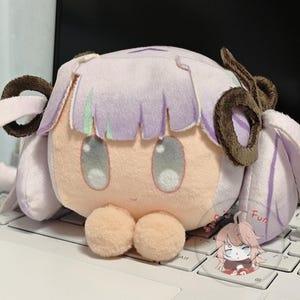 May include: A plush doll with a light purple head, brown ear-like accents, and large gray eyes. The doll has a peach-colored face and two peach-colored spheres at the bottom. A small illustration of a character is on the keyboard.