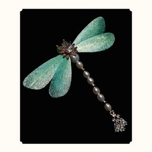 May include: A dragonfly brooch with teal wings and a body of pearls and gold beads. The head features a flower design with a central crystal. The dragonfly's tail has a cluster of crystals.