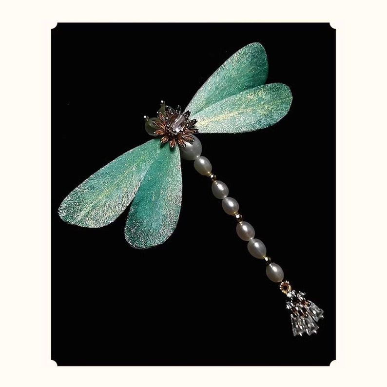 May include: A dragonfly brooch with teal wings and a body of pearls and gold beads. The head features a flower design with a central crystal. The dragonfly's tail has a cluster of crystals.