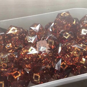 May include: A close-up of a container filled with many brown and gold dice. The dice are made of clear glass with gold flecks and have a geometric design.