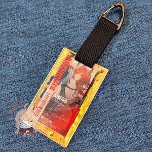 May include: A clear plastic ID holder with a black strap and metal clip. The holder features a colorful anime graphic with the text "Arknights" and "Laevatain". The holder is rectangular with a yellow border and a red background.