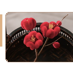 May include: Red artificial flowers with yellow stamens are arranged in a dark, decorative basket. The flowers are in full bloom, with a soft, velvety texture. The image includes text in a vertical column.