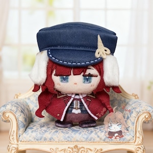 May include: A plush doll with red hair, a navy blue cap, and a red jacket. The doll has blue eyes and is sitting on a small, ornate chair with a blue floral pattern. A small illustration is attached to the chair.