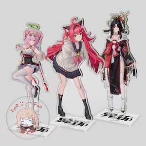 Puede incluir: Tres figuras de acrílico con personajes de estilo anime. La figura de la izquierda tiene cabello rosa y un halo. La figura central tiene cabello largo rojo y un suéter blanco. La figura de la derecha tiene cabello negro y un traje estilo kimono.