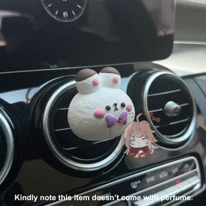 May include: White bunny-shaped car air freshener with a purple bow. The air freshener is attached to a car's air vent. The air freshener is in the shape of a bunny with a brown nose and pink cheeks. The air freshener is in a car with black interior.