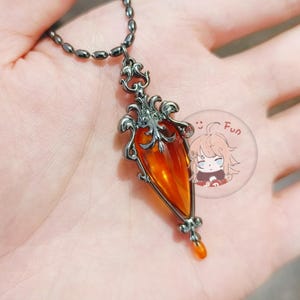 May include: A pendant necklace featuring a teardrop-shaped orange gemstone set in an ornate, dark metal frame. The necklace has a matching dark chain. A small, round sticker with a cartoon character and the word "Fun" is visible.