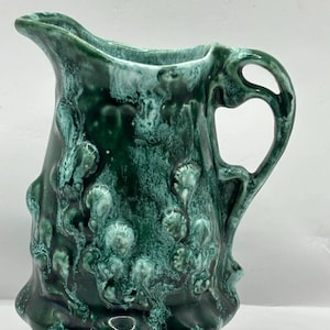 Puede incluir: Una jarra de cerámica verde con un diseño orgánico texturizado. La jarra tiene un pico, un asa curva y una base. El esmalte tiene un aspecto moteado con tonos de verde oscuro y claro.