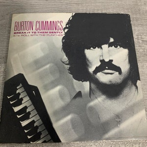 May include: A black and white photo of Burton Cummings, a Canadian musician, on a record cover. The text on the cover reads "Burton Cummings, Break It To Them Gently, b/w Roll With The Punches".