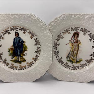 Puede incluir: Dos platos cuadrados de cerámica blanca con ribetes dorados y un diseño en relieve. Cada plato presenta una imagen diferente de una persona con ropa de época. Un plato presenta a un hombre con un traje azul y el otro a una mujer con un vestido amarillo.