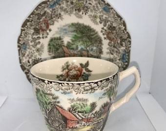 May include: A vintage teacup and saucer set with a brown, green, and red floral pattern. The pattern features a country scene with a red barn, trees, and a stream.