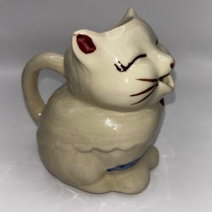 Peut inclure: Un pot à crème en céramique blanche en forme de chat avec un motif bleu et blanc sur le fond. Le chat a un nez rouge et des moustaches brunes.