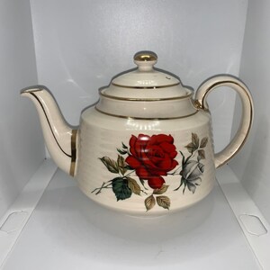 May include: A white teapot with a gold trim and a red and white rose design.