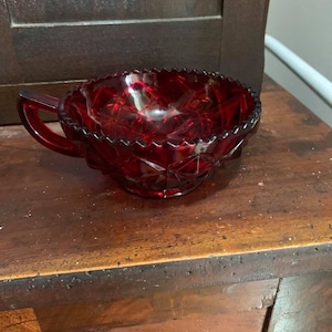 May include: A ruby red glass creamer with a handle and a decorative, scalloped rim. The creamer has a diamond-cut pattern and sits on a wooden surface. The antique creamer is a vintage piece.