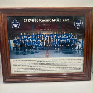 Könnte beinhalten: Ein gerahmtes Foto des Eishockeyteams Toronto Maple Leafs von 1997-1998 in einem Holzrahmen. Das Bild zeigt das Team auf dem Eis, mit dem Text 1997-1998 Toronto Maple Leafs oben und einer Liste der Spieler- und Mitarbeiternamen darunter.