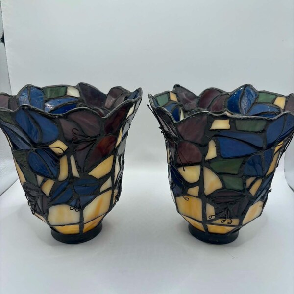 May include: Two stained glass lamp shades. Each shade features a floral design with blue, purple, green, and yellow glass pieces. The shades have a scalloped edge and a black base. The shades are approximately 6 inches tall.