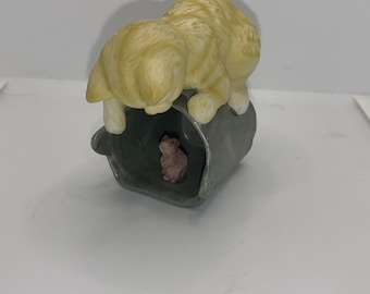 May include: A ceramic figurine of a yellow tabby cat peering into a gray bucket. Inside the bucket is a small pink mouse. The cat is in a crouching position, with its head and front paws over the bucket.
