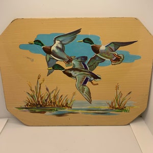 May include: A wooden plaque featuring a painted image of three mallard ducks in flight over a blue lake. The ducks are depicted in shades of green, brown, and blue. The lake is surrounded by green reeds.