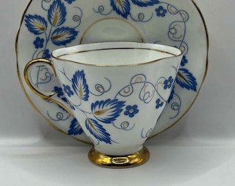 May include: White teacup and saucer set with blue floral and gold trim. The teacup has a gold base and handle. The saucer has a matching design. The set is likely vintage or antique.
