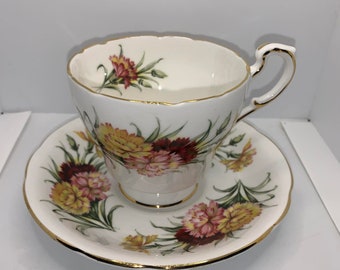 May include: A white teacup and saucer set with a floral pattern of pink and yellow carnations. The set has a gold trim around the edges.