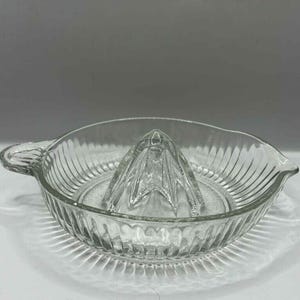 Peut inclure: Presse-agrumes en verre transparent. Il comporte un bol rainuré, un presse-agrumes central, une poignée et un bec verseur. Le design est simple et fonctionnel, adapté à une utilisation en cuisine.