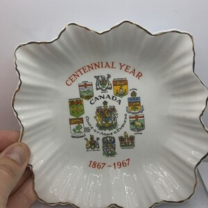 Puede incluir: Plato de cerámica blanca con borde dorado y un diseño rojo y negro que celebra el centenario de Canadá, 1867-1967. El plato presenta los escudos de armas y los emblemas de las diez provincias.