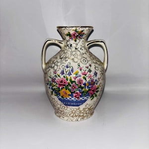 Puede incluir: Jarrón color crema con dos asas y diseño floral. El jarrón presenta un arreglo floral estilo punto de cruz en una cesta azul. Ribetes dorados acentúan el borde y las asas. El jarrón tiene un patrón texturizado y punteado.