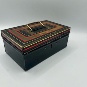 Peut inclure: Une boîte à argent vintage en métal noir avec un couvercle décoratif orné de touches de rouge et d'or. La boîte est dotée d'une poignée en laiton et d'une serrure sur le devant. Cette boîte rectangulaire est probablement utilisée pour ranger de l'argent ou des objets de valeur.