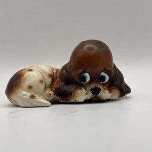 May include: A ceramic figurine of a sad-looking puppy, lying down with its head resting on its paws. The puppy has brown fur on its head and ears, with white and brown spots on its body. It has big blue eyes and a black nose.