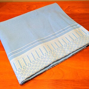 Pode incluir: Têxtil azul claro dobrado com uma borda branca estampada. A borda apresenta um design geométrico de diamantes e linhas verticais. O têxtil é provavelmente um cobertor ou um tecido decorativo.