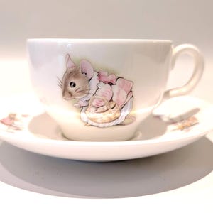 May include: A white ceramic teacup and saucer set featuring a detailed illustration of a brown mouse wearing a pink outfit and carrying a small woven basket. The mouse is depicted in a soft, watercolor style on the side of the cup and saucer.