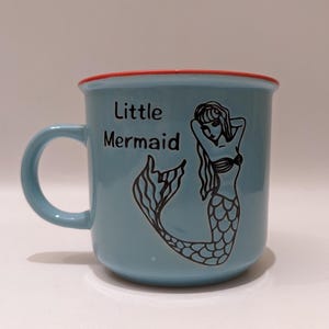 May include: A light blue ceramic mug with a red rim. The mug features a black and white illustration of a mermaid with the text "Little Mermaid" printed on the front.