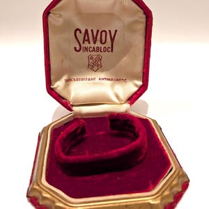 May include: Open red velvet jewelry box with gold trim. The inside is lined with cream-colored satin and features the text "SAVOY INCABLOC SHOCKRESISTANT ANTIMAGNETIC". The box is designed to hold a bracelet or watch.