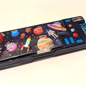Peut inclure: Trousse noire avec un thème spatial. Le couvercle présente des planètes, une fusée et un astronaute sur un fond étoilé. La trousse a une calculatrice intégrée et des boutons bleus. La trousse est rectangulaire et possède un fermoir argenté.