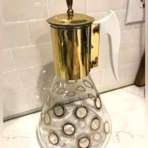 May include: A vintage glass and brass coffee pot with a white handle. The clear glass body features a pattern of gold circles. The top and handle are made of shiny brass. The coffee pot is a decorative kitchen item.