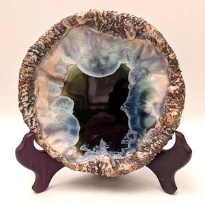 May include: A ceramic geode sculpture with a dark interior and a textured, brown and beige outer edge. The interior features blue, green, and white crystalline formations. The sculpture is displayed on a dark purple wooden stand.
