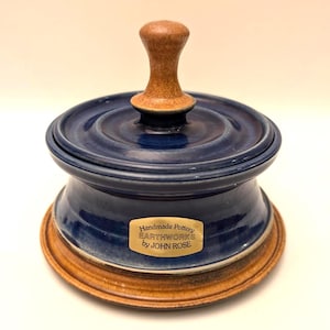 Peut inclure: Beurrier en poterie fait à la main, corps émaillé bleu marine et poignée de couvercle en bois naturel. La base est de couleur marron chaud. Le plat porte une étiquette indiquant "Handmade Pottery EARTHWORKS by JOHN ROSE."