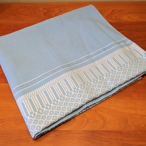 Peut inclure: Une nappe bleu clair avec une bordure brodée blanche. La bordure présente un motif répété de diamants et de lignes verticales.