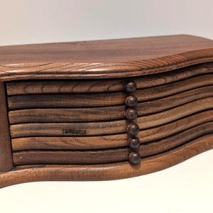 May include: A wooden coaster set with a dark brown finish. The set includes a curved top and base, with multiple wavy, horizontal coasters. Six small, round, dark brown knobs are on the side. The coasters are made of wood.